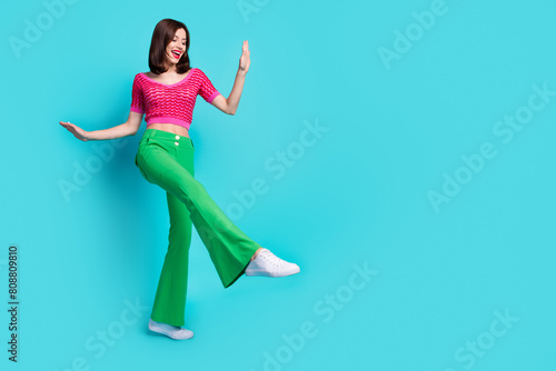 Full size photo of positive girl wear pink knit top green trousers dancing look empty space show shoe isolated on blue color background