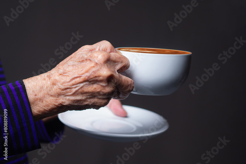 Close-up of a senior woman's hand holding a white coffee cup while standing on a gray background.
