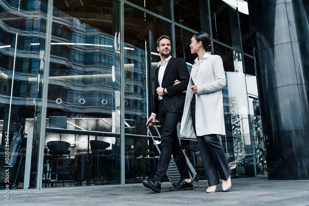 Fototapeta premium Business couple in coats walking out the airport with suitcase during the business trip. Businessman and businesswoman talking and hold luggage travel to business trip.