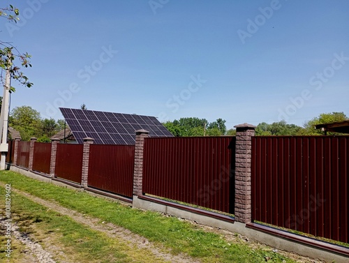 Solar panels are mounted behind a tall red sheet metal fence to generate solar energy into electrical energy. The topic of using solar energy for human needs. Electricity generation from renewable nat