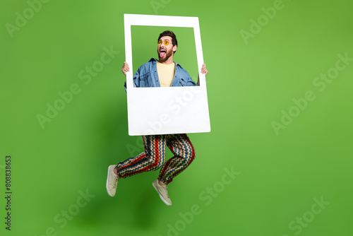 Full length photo of impressed excited guy dressed jeans shirt jumping high holding photo frame isolated green color background