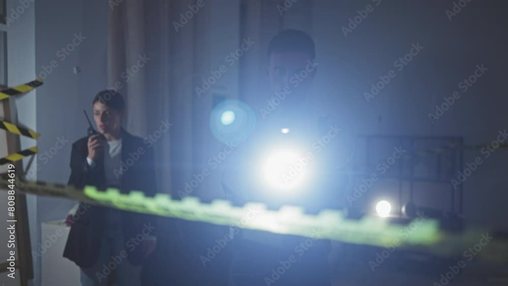 Investigators, a man and woman, search a dimly lit indoor crime scene with caution tape and flashlights. Investigators, a man and woman, search a dimly lit indoor crime scene with caution tape and flashlights.