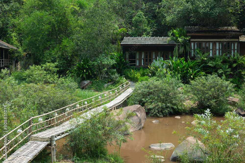 In the natural scenery of Sapan Village, Nan Province, a bamboo bridge crosses a rocky river amidst resorts along the shore, its murky waters adding allure.