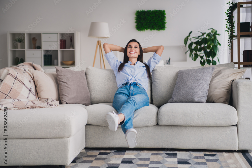 Full length portrait of pretty lady hands behind head sit sofa wear shirt bright interior apartment indoors Full length portrait of pretty lady hands behind head sit sofa wear shirt bright interior apartment indoors