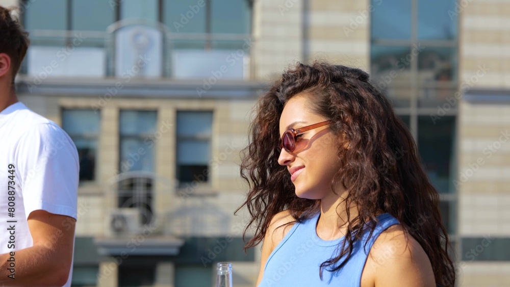 Close up of pretty attractive young Caucasian woman with curly hair in sunglasses dancing with bottle of beer on sunny day outdoor at a party. People dance on background. Slow motion