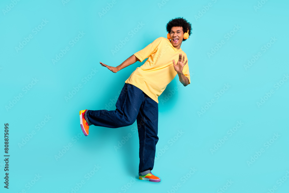 Full size photo of carefree man wear oversize t-shirt denim pants dancing in headphones have fun isolated on teal color background