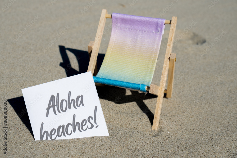 ALOHA BEACHES text on paper greeting card on background of beach chair lounge summer vacation decor. Sandy beach sun. Holiday concept postcard. Travel Business concept ALOHA BEACHES text on paper greeting card on background of beach chair lounge summer vacation decor. Sandy beach sun. Holiday concept postcard. Travel Business concept