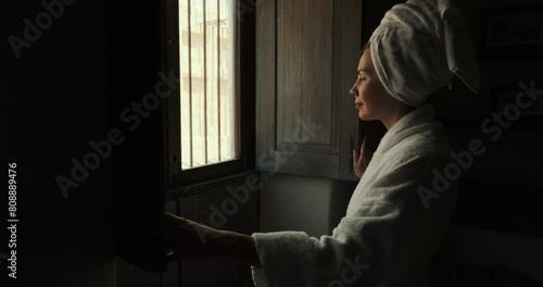 Woman in dark room old hotel standing in robe. Young leady in bathrobe and with towel on her head, looking in window and smiling. Girl has opened the shutters and is standing looking out window photo