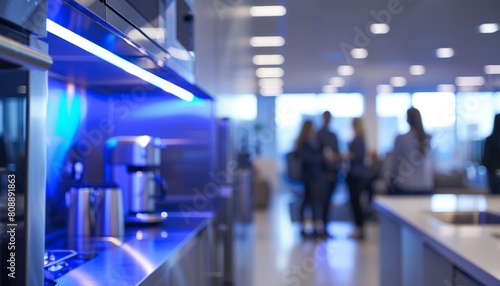 A minimalist office kitchen with sleek stainless steel appliances and blue LED lighting The background is a soft blur of people grabbing coffee and socializing