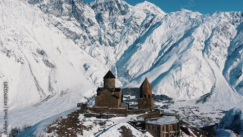 Aerial view of ancient stone churche perched atop a ridge, surrounded by the vast, snow-covered mountains under a clear blue sky.  photo