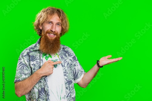 Young bearded man showing thumbs up and pointing right empty place, advertising area for commercial text copy space for goods promotion advertisement. Guy isolated on chroma key background. Lifestyles