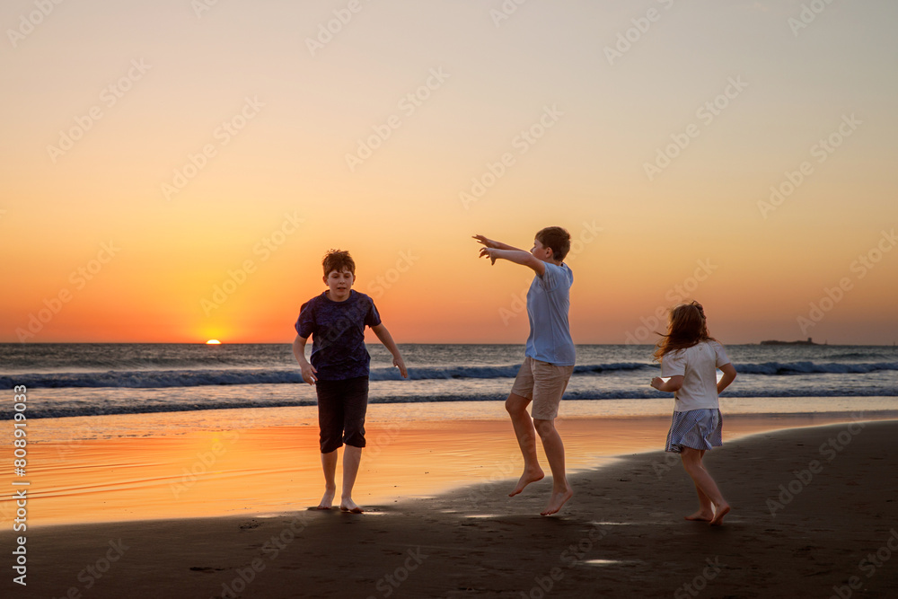 Three kids silhouettes running and jumping on beach at sunset. happy family, two school boys and one little preschool girl. Siblings having fun together. Bonding and family vacation.