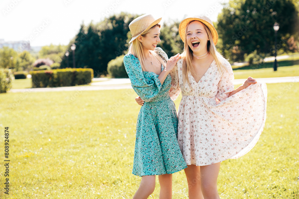 Two young beautiful smiling hipster woman in trendy summer dress. Carefree women in the street in hats. Positive models at sunset. Cheerful and happy. Walking in grass, enjoy summertime days