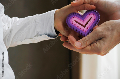 Close-up of the hand of a Caucasian boy dressed in a white shirt giving a purple pink heart made of ironing beads into the hands of a Caucasian woman. Mother's Day concept with copy space.