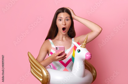amazed excited teen girl with open mouth and inflatable unicorn ring hold phone in one hand and hold head with the other, young blogger, shocking news wear colourful swimsuit pink background isolated