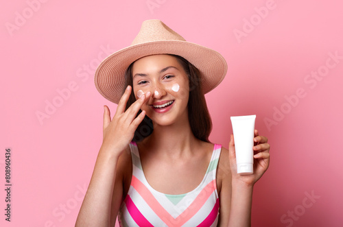 Cheerful smiling tanned teen girl put sunscreen on her face and holds a white blank cream tube mock up in hand, wear swimsuit and straw hat isolated on pink background sun protection skin care concept