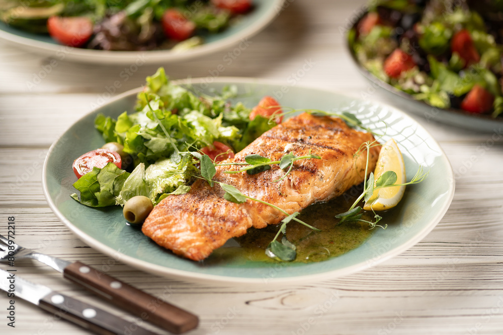 One piece of salmon grilled with lettuce and vegetables salad. Serving dishes on table One piece of salmon grilled with lettuce and vegetables salad. Serving dishes on table