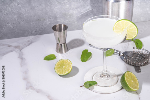 Gimlet citrus alcohol cocktail. Sweet and sour boozy alcoholic drink, with gin or vodka and lime garnish, on light grey with hard light and bar utensils