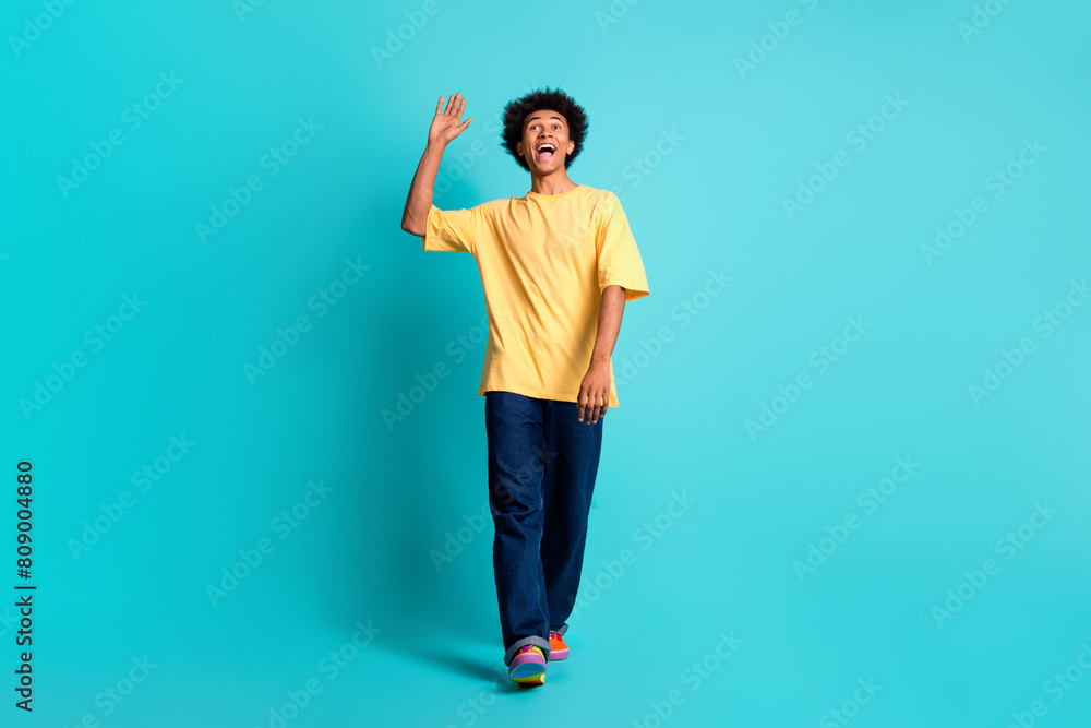 Full size photo of nice man wear oversize t-shirt denim pants look at friend in empty space waving hand isolated on teal color background Full size photo of nice man wear oversize t-shirt denim pants look at friend in empty space waving hand isolated on teal color background