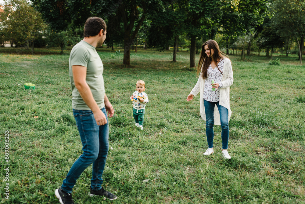Fototapeta premium Happy mother, father hug baby son walking in garden at sunset. Family spending time together outdoors. Child with parents happiness playing in green grass in park. Children's day. Friendly family.