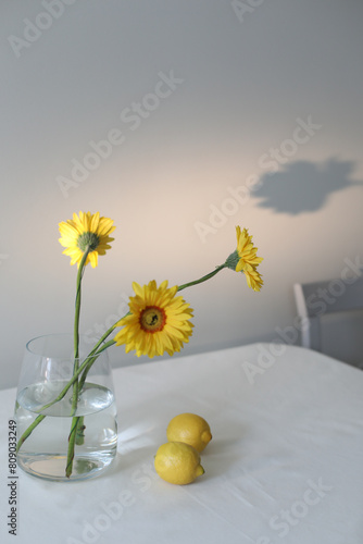 still life with lemons and yellow gerberas in a glass jar on a table covered with linen tablecloth. Summer composition with lemons and yellow flowers on kitchen table.
