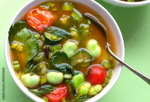 Spring minestrone with fave beans, tomatoes, celery, carrots and aromatic herbs, spring vegetables, vegetarian proteins on a green mint background. Italian cuisine, fresh traditional Easter ingredient