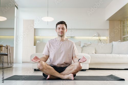 A man sits cross-legged on a yoga mat, meditating peacefully in a lotus position within a bright and modern living space, embodying tranquility and mindfulness.