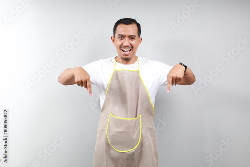 Friendly asian barista in brown apron pointing fingers down, showing your logo banner, standing over white background photo