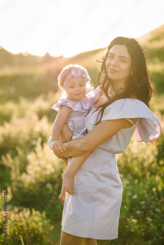 Fototapeta premium Young mother kissing baby girl walk in park. Family holiday in garden. Portrait mom with child together on nature. Mum, little daughter outdoors. Happy Mothers Day. Close up.