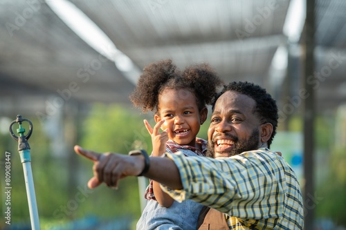 Happy african black parents dad father with daughter child on piggyback teasing fun in garden greenhouse. Black daughter kiss cheek dad and neck riding in vegetable greenhouse garden in light sunset photo