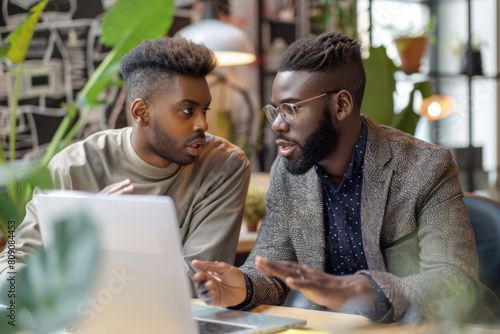 Two business people discussing in office setting. Team leader sits beside new team member at workplace and explains project details to new colleague