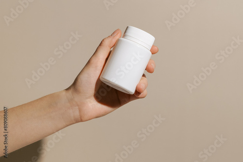 Female hand holding a white mockup jar with dietary supplement capsules on a beige background. Concept of pharmacy and medicine, health care