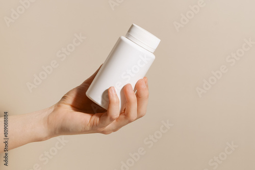 Female hand holding white plastic mockup pill jar on beige isolated background. Concept of vitamins for beauty and health, dietary supplements and pharmacy.