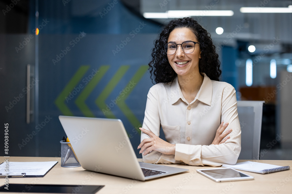 A cheerful Hispanic businesswoman with glasses smiling confidently at her desk in a modern office. She is surrounded by digital devices, showcasing professionalism and positivity. A cheerful Hispanic businesswoman with glasses smiling confidently at her desk in a modern office. She is surrounded by digital devices, showcasing professionalism and positivity.