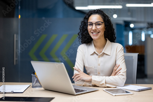 A cheerful Hispanic businesswoman with glasses smiling confidently at her desk in a modern office. She is surrounded by digital devices, showcasing professionalism and positivity.