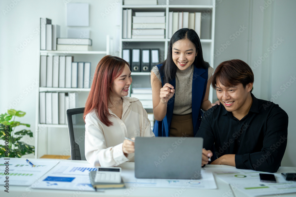 Three people are sitting at a desk with a laptop open. They are smiling and seem to be having a good time Three people are sitting at a desk with a laptop open. They are smiling and seem to be having a good time