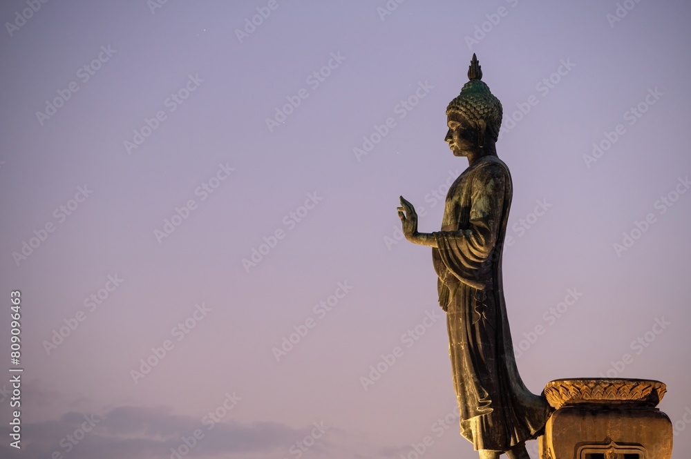 Fototapeta premium Stand big Buddha Statue in silhouette sun set Light background in park of Thailand temple.Yellow orange light silhouette dark shadow of image Buddha statue stand Buddha tall walk in sun light cloud.