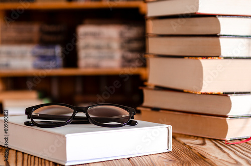 Book stacked on Laptop with glasses on wooden Table with blurred Bookshelf in library room