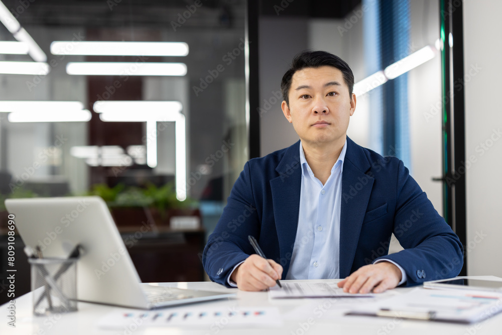 Focused Asian businessman in a well-fitted blue suit working at his office desk surrounded by documents and a laptop, exuding professionalism and determination. Focused Asian businessman in a well-fitted blue suit working at his office desk surrounded by documents and a laptop, exuding professionalism and determination.