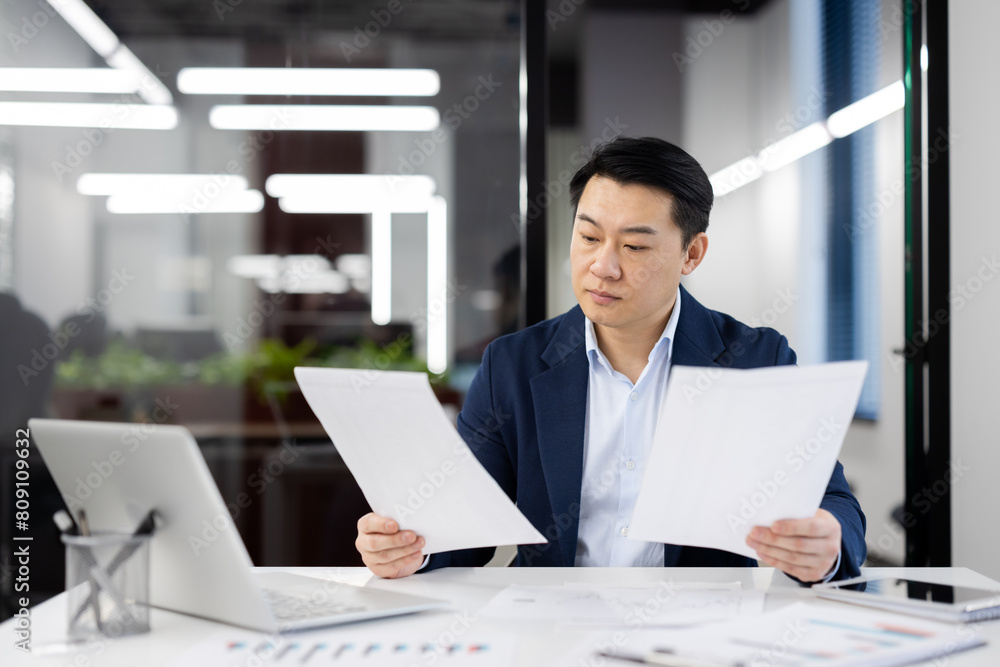 Focused Asian businessman in a blue suit reviews paperwork while working at his desk in a well-lit modern office. Concept of business analysis and professional diligence. Focused Asian businessman in a blue suit reviews paperwork while working at his desk in a well-lit modern office. Concept of business analysis and professional diligence.