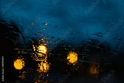 vista macro sulla superficie di vetro di un'auto bagnata dalla pioggia durante un temporale serale con sfondo sfuocato e con sfumature di blu e nero e alcune luci arancioni photo