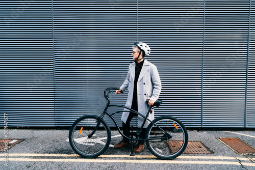 Stylish male in coat, sunglasses and protective helmet standing with retro bicycle near gray urban wall. Neutral carbon footprint transportation. Green eco friendly mobility sustainable transport