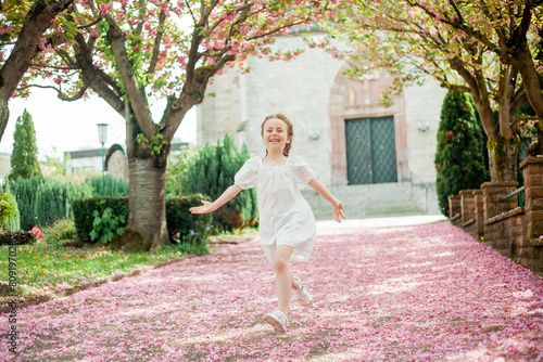 Happy pretty girl 6 years old in white dress near blooming pink sakura. The child is walking in the park. Spring.