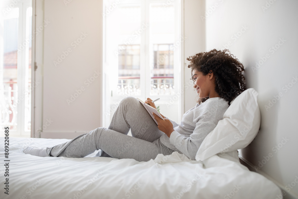 Hispanic woman is seated on a bed, focused on writing in a notebook. She appears to be deep in thought, jotting down ideas or reflections. The room is simple, with neutral decor and soft lighting. Hispanic woman is seated on a bed, focused on writing in a notebook. She appears to be deep in thought, jotting down ideas or reflections. The room is simple, with neutral decor and soft lighting.