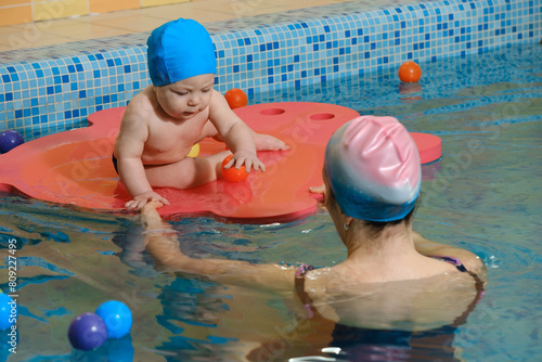 Early age swimming in pool. Baby boy trained to swim in water. Happy child with trainer woman in indoor swimming pool playing and having fun. Healthy and sport family with infant, active parent