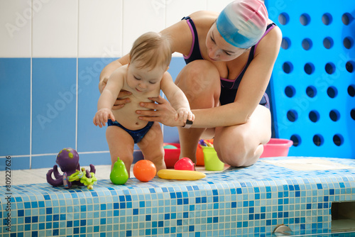 Early age swimming in pool. Baby boy waiting near side, learning shapes and fruits. Happy child with coach woman in indoor swimming pool playing and having fun. Healthy and sport family with infant