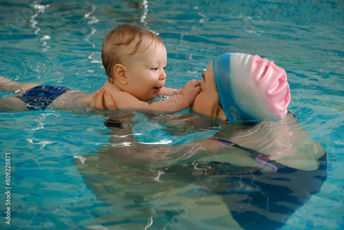 Early age swimming in pool. Baby boy trained to swim in water. Happy child with trainer woman in indoor swimming pool playing and having fun. Healthy and sport family with infant, active parent