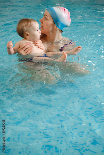 Early age swimming in pool. Baby boy trained to swim in water. Happy child with trainer woman in indoor swimming pool playing and having fun. Healthy and sport family with infant, active parent