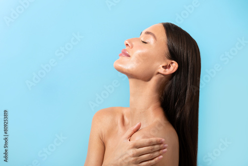 Charming pretty brown-haired lady hug her naked shoulders, posing with closed eyes isolated on blue background with copy space