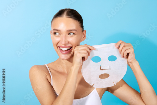 Attractive European woman holding paper sheet mask, enjoys beauty skin care, smiling positively, standing isolated on blue background photo